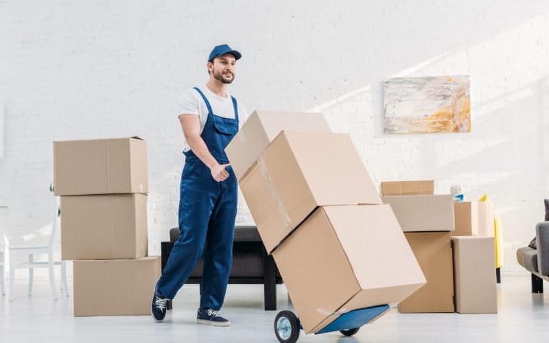 mover in uniform transporting  cardboard boxes on hand truck in apartment